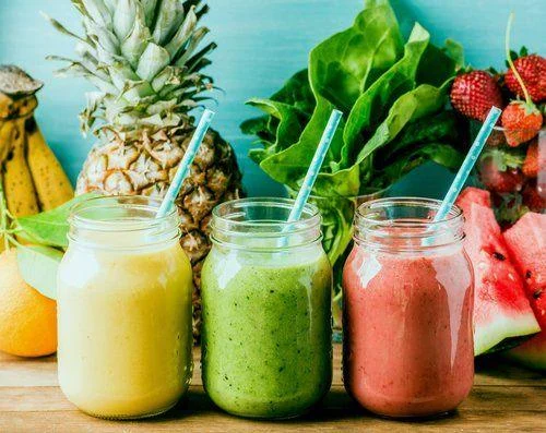GettyImages-543809310 Three mason jars filled with yellow, green, and pink smoothies with straws, surrounded by assorted fresh fruits and leafy greens on a wooden surface.