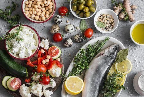 Mediterranean style food background. Fish, vegetables, herbs, chickpeas, olives, cheese on grey background, top view. Healthy food concept. Flat lay A whole fish on a platter with lemon and herbs, surrounded by bowls of chickpeas, olives, cheese, vegetables, eggs, and a dish of oil on a gray surface.