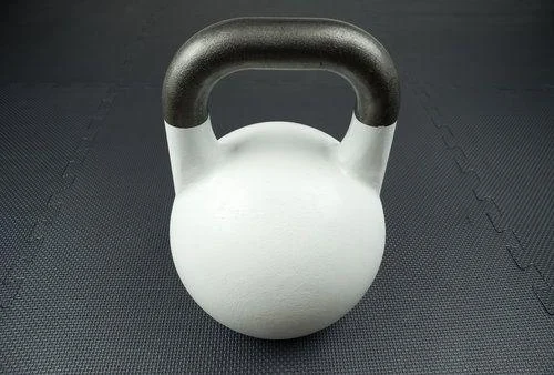 White competition kettlebell on a gym floor A white kettlebell with a black handle sits on a textured dark gym floor.