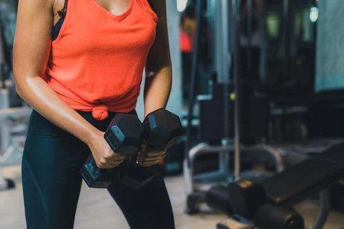 Heavy weight exercise Person in an orange tank top and black leggings holding two dumbbells at a gym, with exercise equipment in the background.
