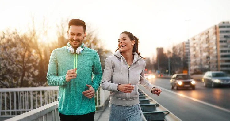 Couple jogging outdoors Two people jogging on a city sidewalk at sunset, both wearing athletic clothing; cars and buildings are visible in the background.