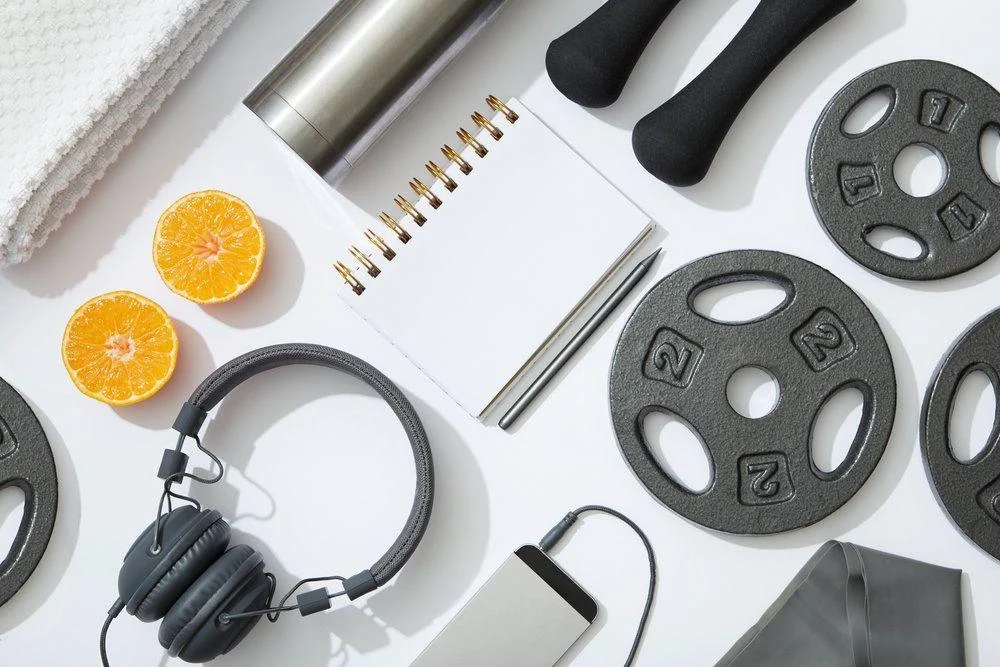Flat lay of fitness equipment including dumbbells, weight plates, a towel, notebook, pen, headphones, resistance band, water bottle, smartphone, and halved orange.