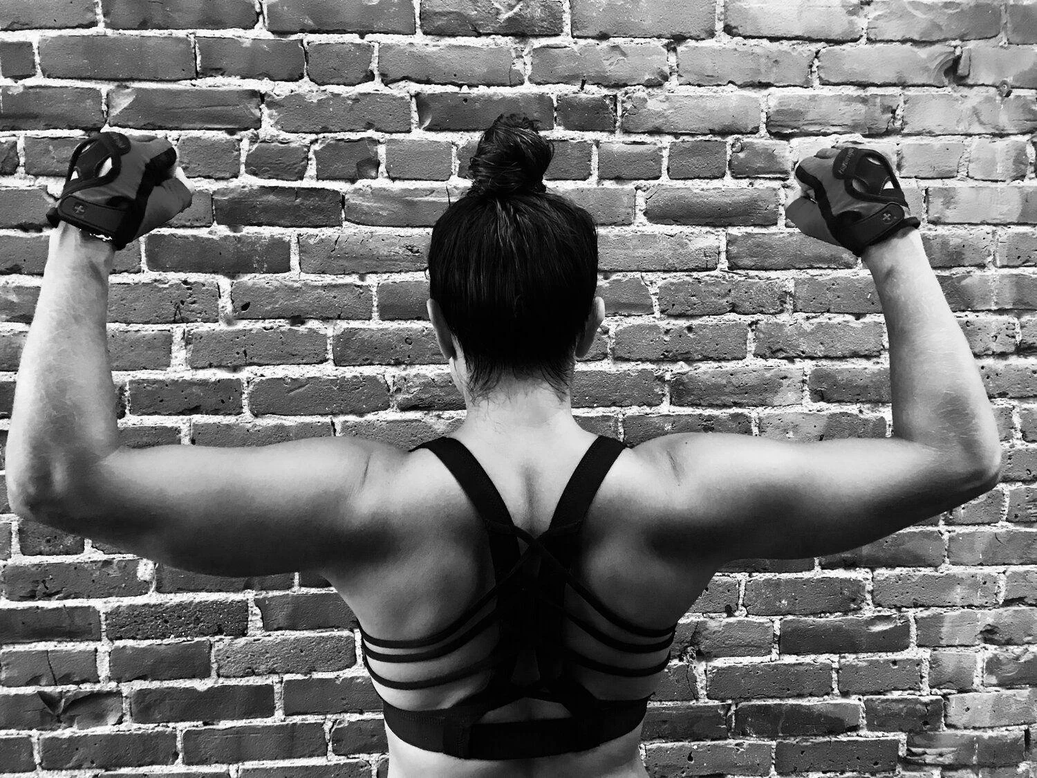 A person faces a brick wall, flexing their arms to show back muscles, wearing athletic gloves and a strappy sports bra.