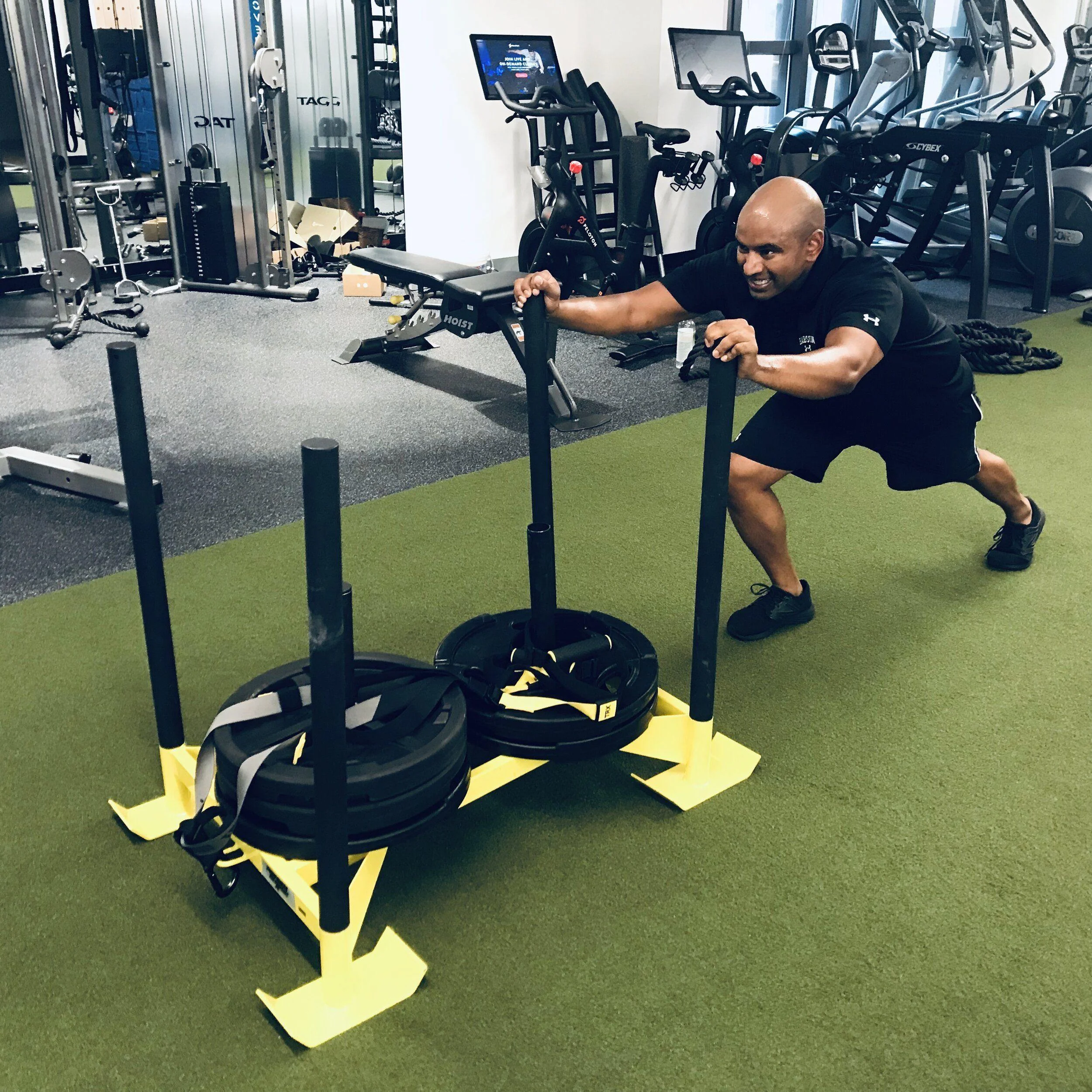 IMG_6642 A man pushes a weighted sled across artificial turf in a gym with various exercise equipment in the background.