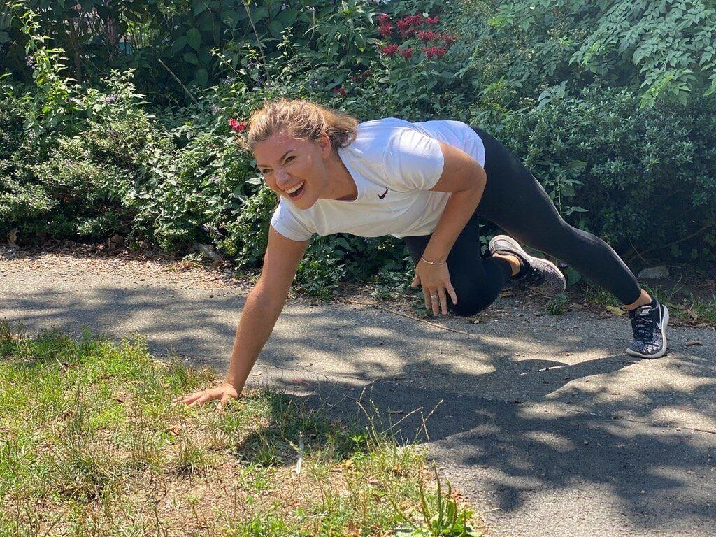 IMG_9141 A woman in athletic wear performs a bear crawl exercise on a paved path outdoors, surrounded by greenery and sunlight.