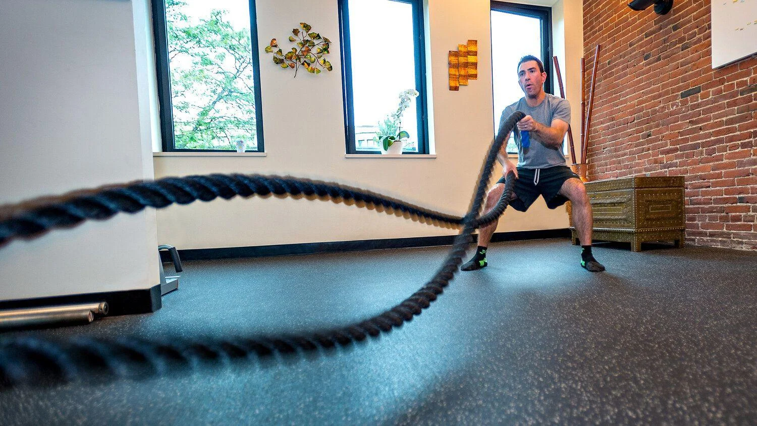 A man exercises indoors, holding and moving battle ropes on a rubber floor in a gym with brick walls and large windows.