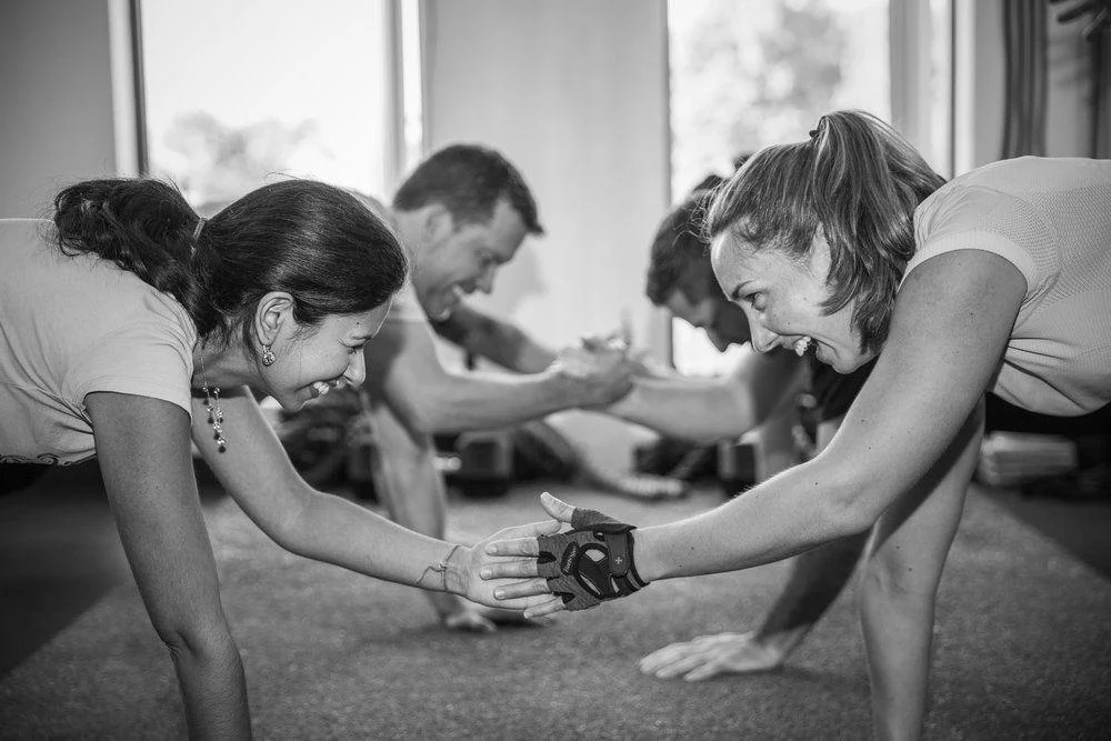 Four people in a gym hold plank positions while reaching out to shake hands and smile at each other.