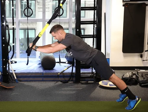 Person performs a suspended plank exercise using TRX straps in a gym, maintaining a straight body position with arms extended overhead.