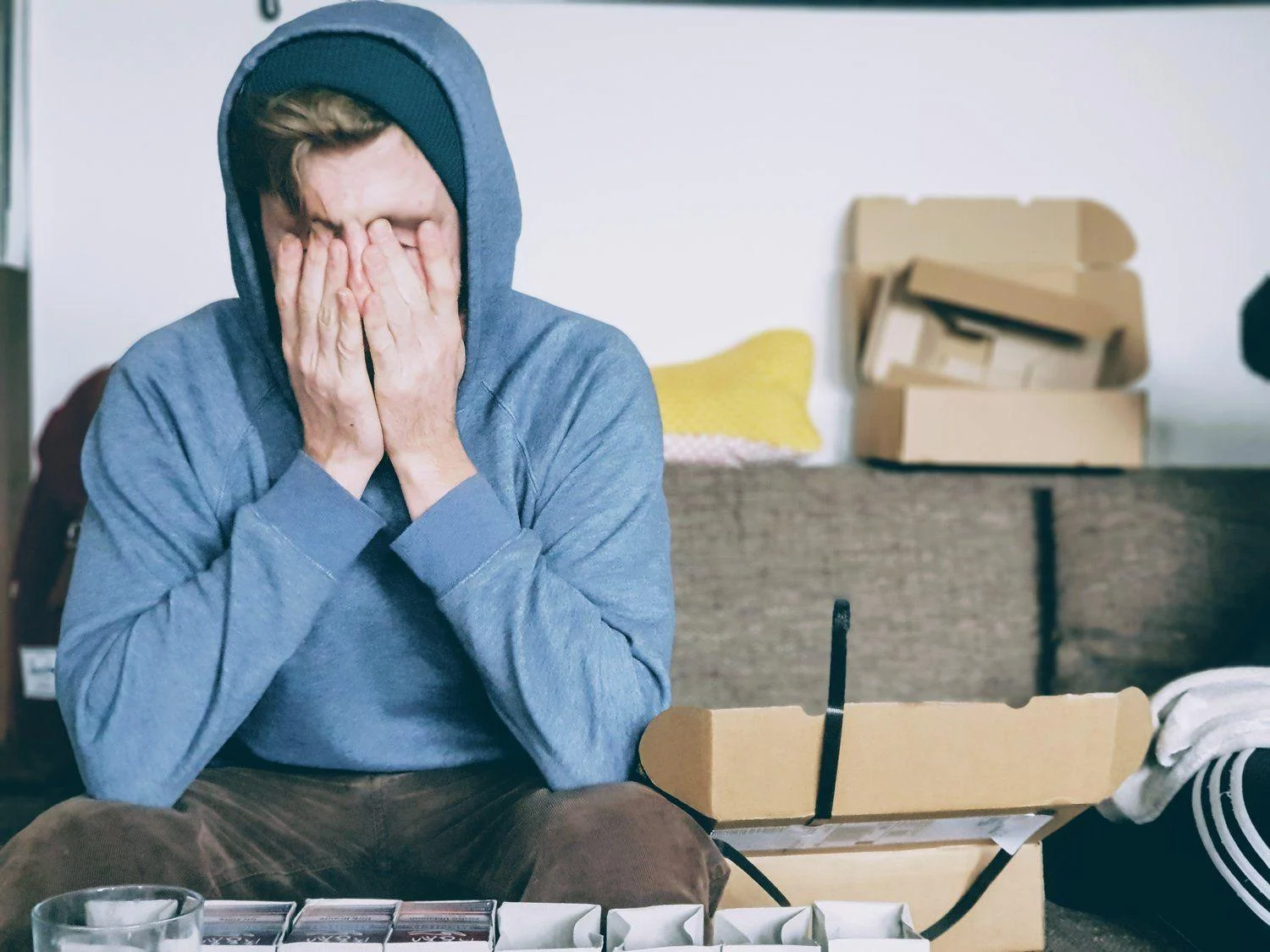 A person in a blue hoodie sits on a couch with their face in their hands, surrounded by open boxes and scattered items.