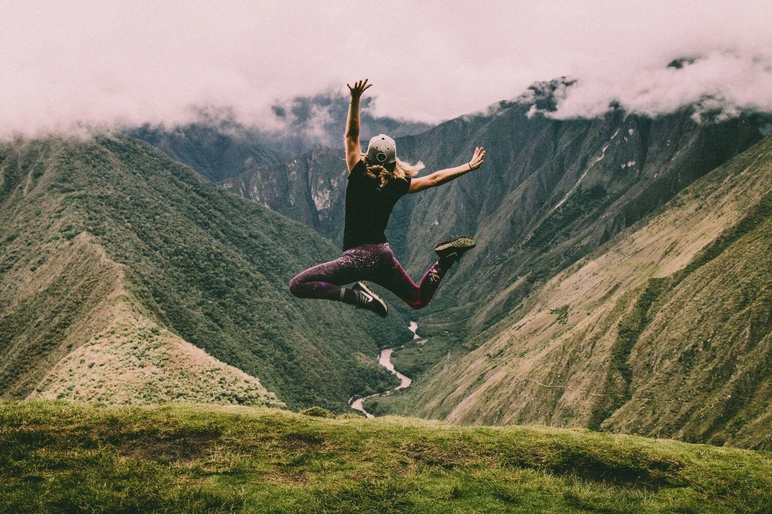 A person wearing a cap and athletic clothes jumps in the air on a grassy hilltop with tall, green mountains and a winding river in the background under cloudy skies.