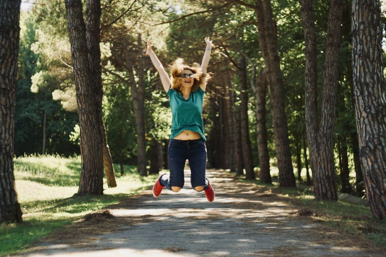 image-asset (8) A person wearing a green shirt and jeans jumps with arms raised on a tree-lined path in a sunlit park.