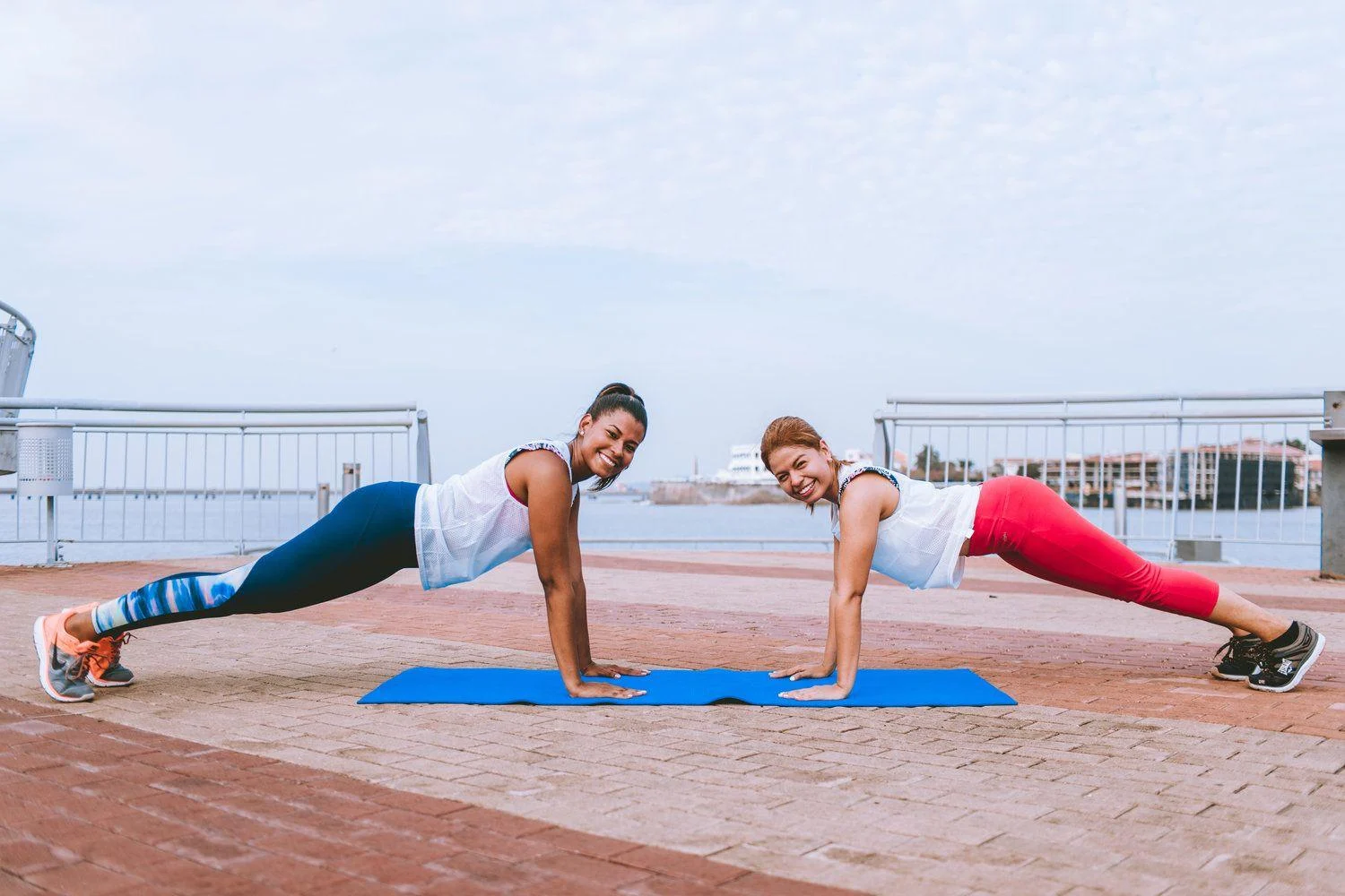 image-asset (9) Two women exercising outdoors on a mat, both holding a plank position and smiling at the camera, with water and buildings in the background.