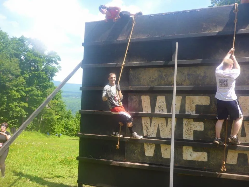 img People climb a large outdoor obstacle wall using ropes on a sunny day; one person sits on the edge, while others ascend or rest on the wall.