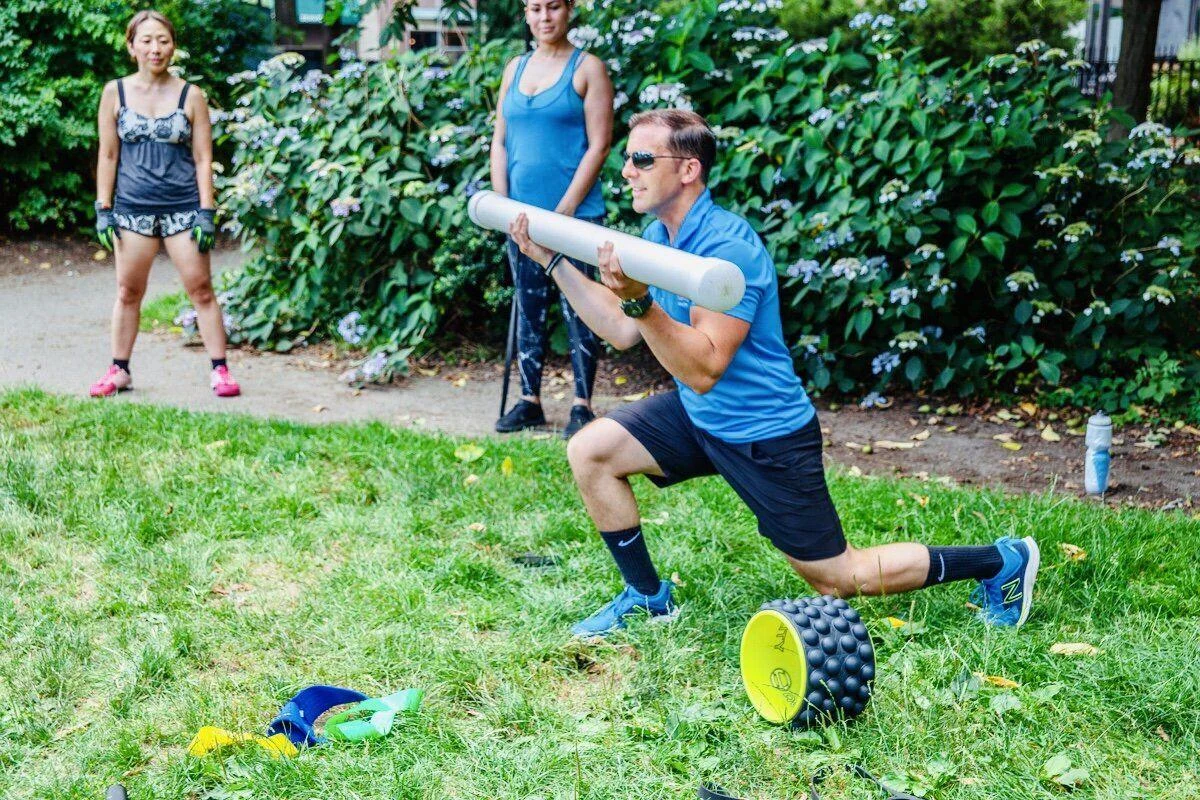 A man in athletic wear does a lunge while holding a weighted tube outdoors, with two women standing and watching in the background. Fitness equipment lies on the grass.