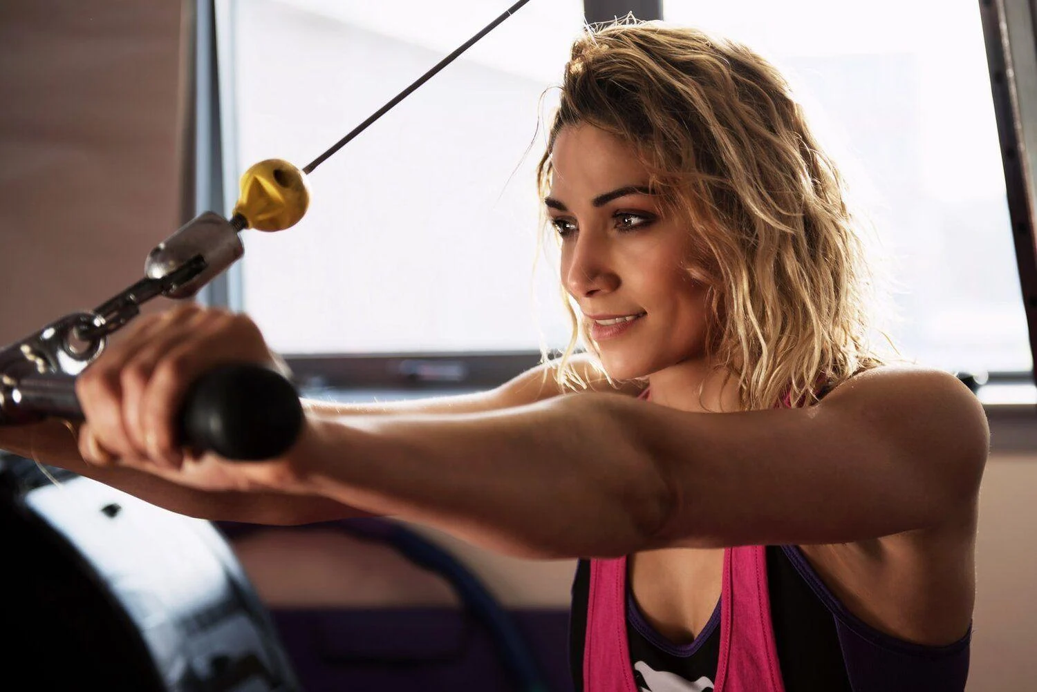 A woman with blonde hair uses a cable machine for a strength training exercise in a gym.