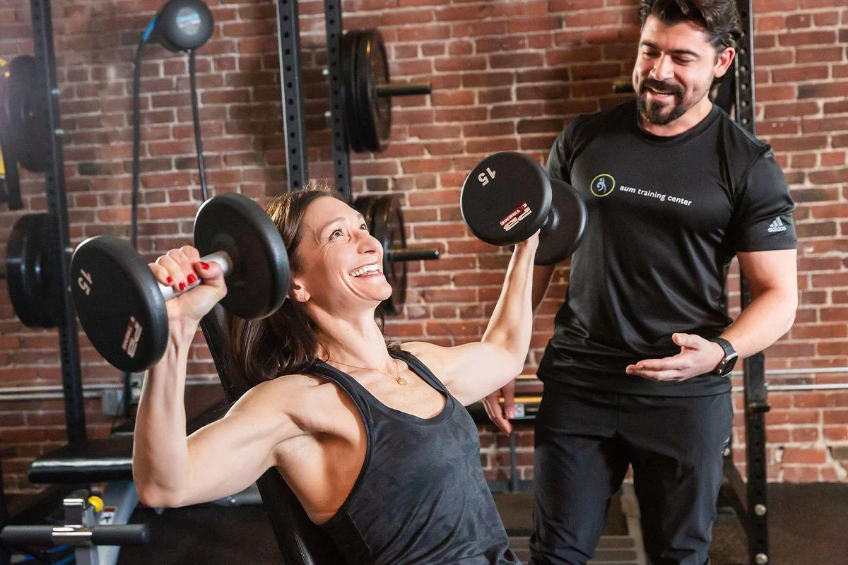 strength-2-1 A woman lifts two dumbbells while seated on a bench, smiling, as a male trainer watches and encourages her in a gym with brick walls.
