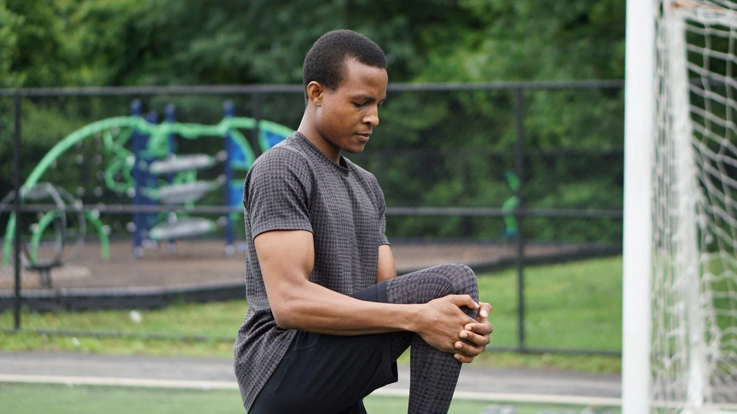 A man in athletic wear does a knee stretch on an outdoor sports field, with playground equipment visible in the background.