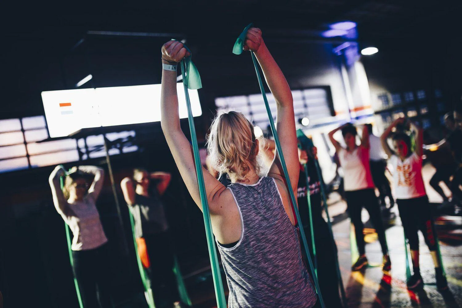 unsplash-image-3RnkZpDqsEI A group fitness class exercises indoors, with participants using resistance bands and raising their arms.