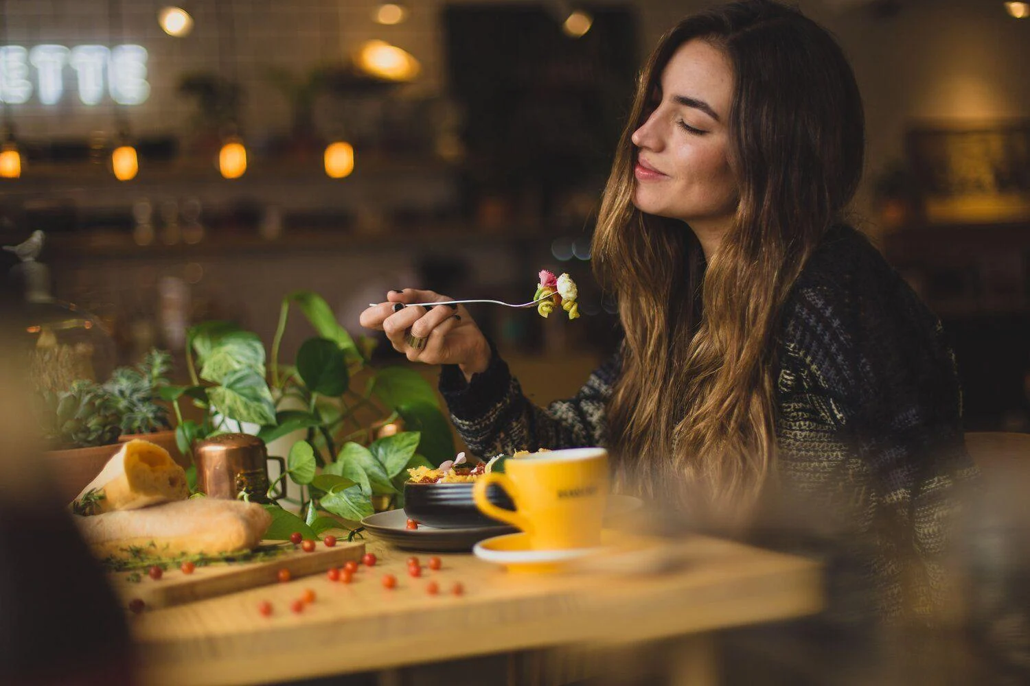 A woman with long brown hair sits at a table in a café, holding a fork with food and smiling, with a yellow cup and various dishes in front of her.