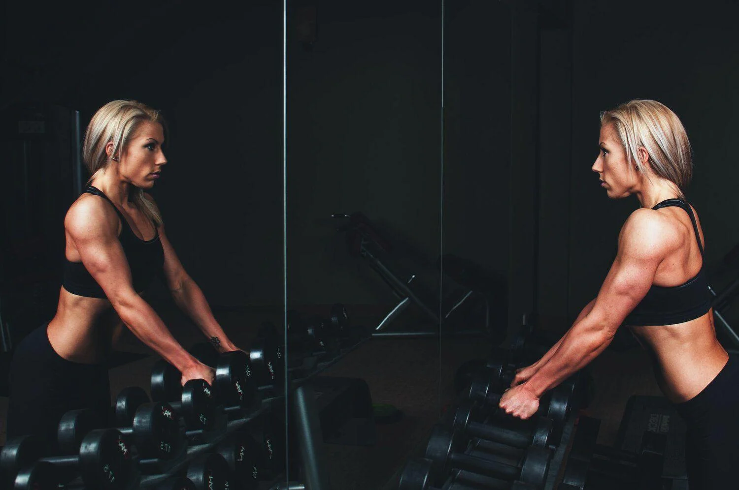 A woman in athletic wear stands in front of a mirror at a gym, gripping dumbbells on a rack and looking at her reflection.