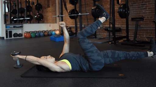 Person lying on an exercise mat in a gym, holding a dumbbell overhead with one arm while raising the opposite leg—a core exercise often included in personal training Boston routines.
