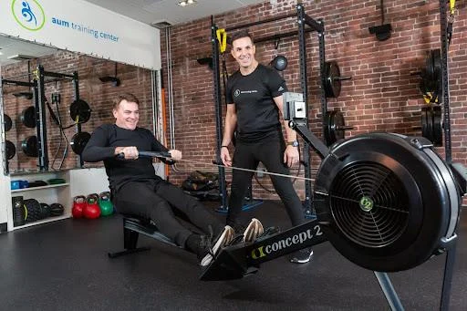 A man uses a rowing machine while another man stands beside him, both in athletic wear, inside a gym with weights and exercise equipment, working together toward their weight loss goals.