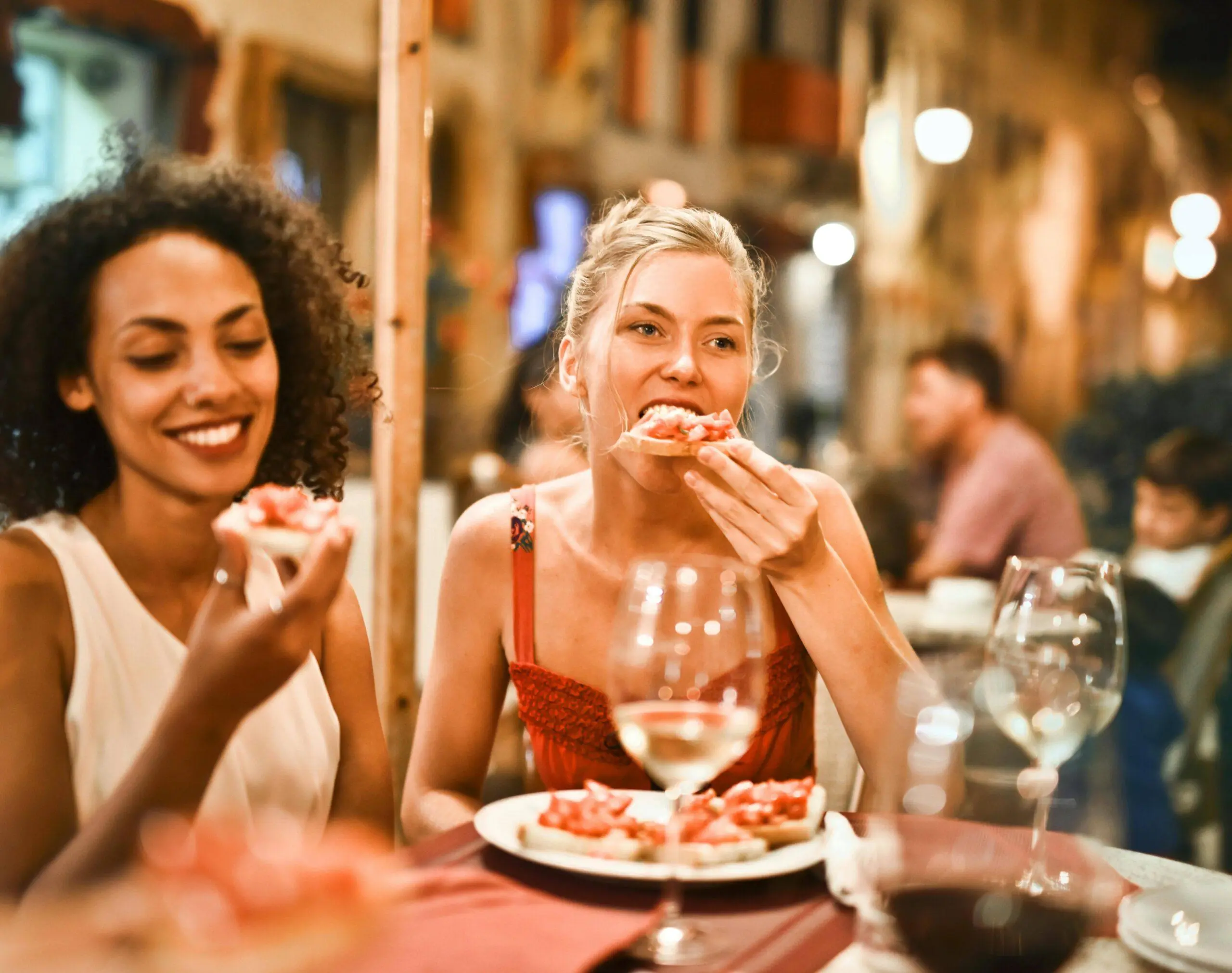 Two women sit at an outdoor restaurant table, enjoying bruschetta and smiling. One chats about her experience with a personal trainer in Boston, while wine glasses and other diners fill the lively background.