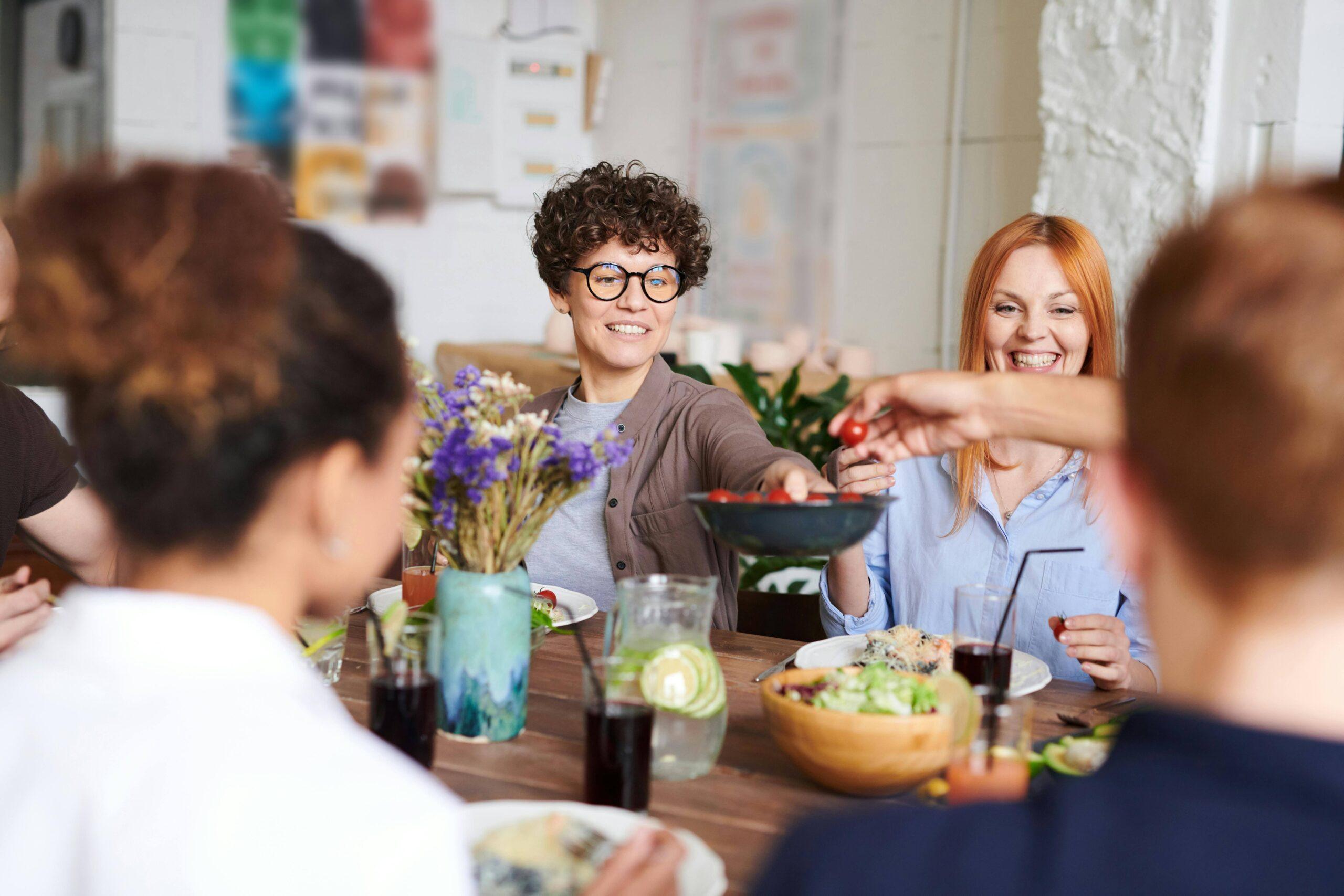 A group of people sit around a table sharing food and drinks, with flowers and pitchers of water in the center, while a Boston personal trainer offers tips for healthy eating.