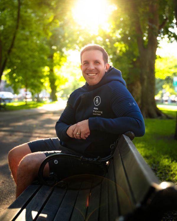A man wearing a navy hoodie sits on a park bench, smiling, with sunlight filtering through green trees in the background. Book a Private Fitness Blueprint Session to start your wellness journey today.