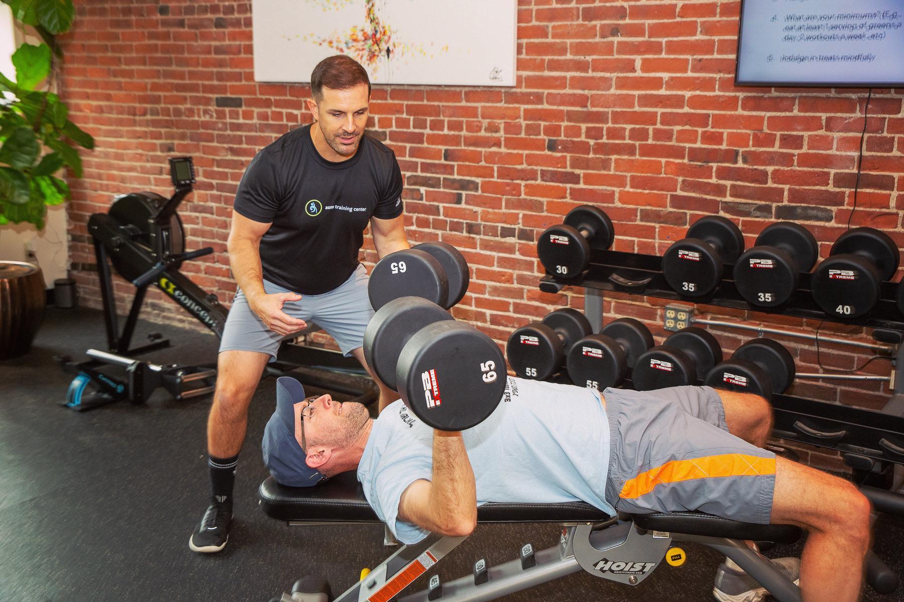 A man lies on a bench lifting dumbbells while another man stands nearby spotting him in a gym with exposed brick walls and various equipment.