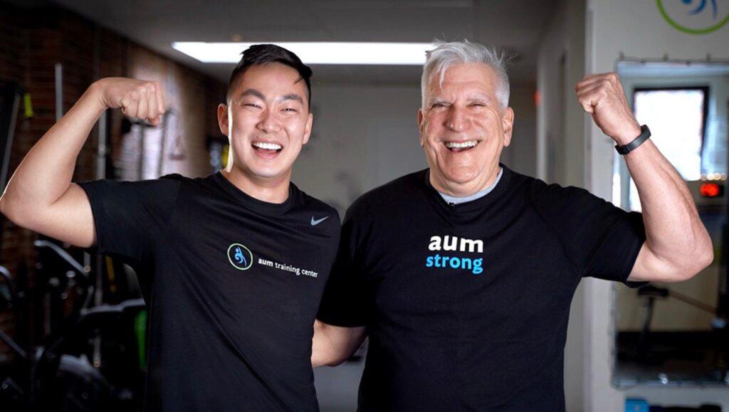 adults-40-get-stronger Two men in workout shirts smile and flex their arms in a gym setting, standing close together with arms around each other's shoulders—showcasing How Adults 40+ Get Stronger through camaraderie and dedication.
