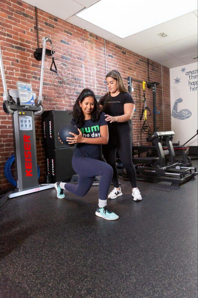 A woman performs a lunge with a medicine ball while another woman, possibly her trainer, assists her in a gym with brick walls and various exercise equipment, showcasing women training together.