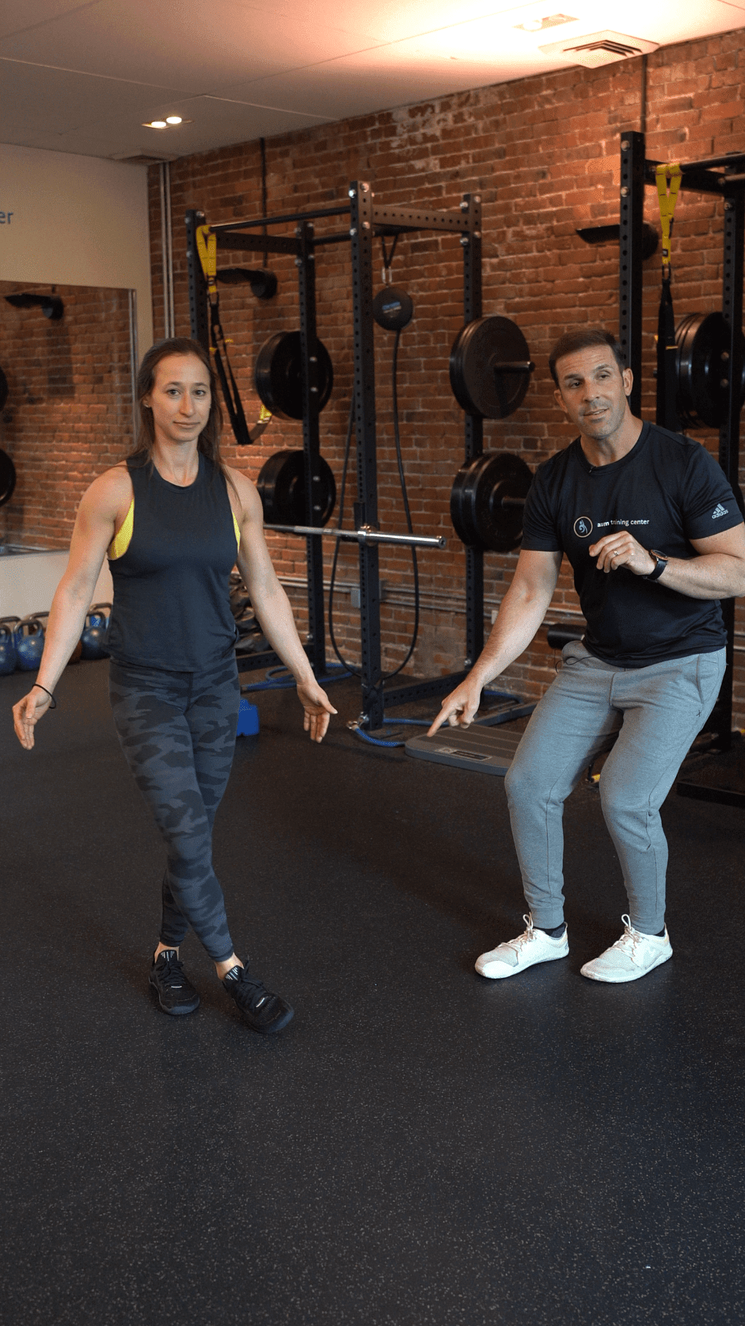 A woman balances on one leg while a man in athletic wear, possibly a Boston personal training coach, gestures toward her in a gym with weights and brick walls in the background.
