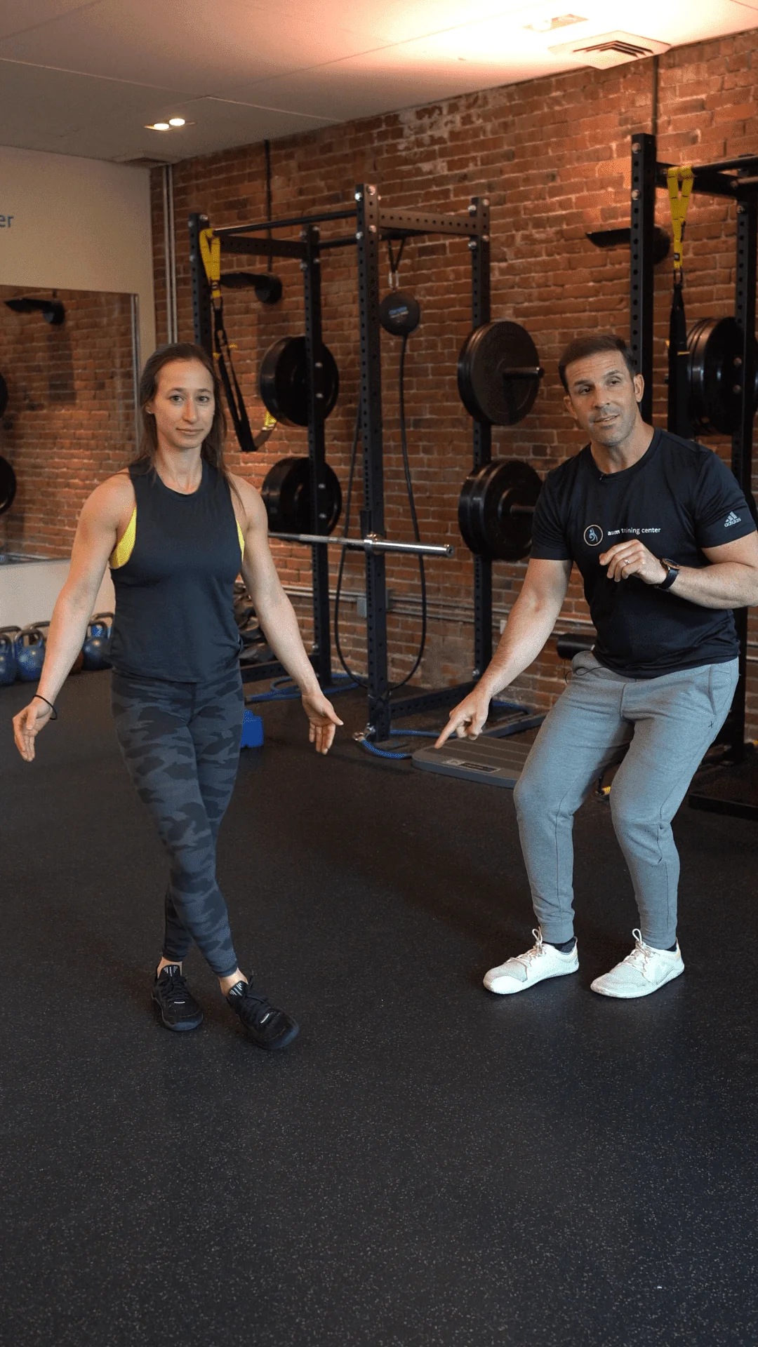 A woman balances on one leg while a man in athletic wear, possibly a Boston personal training coach, gestures toward her in a gym with weights and brick walls in the background.