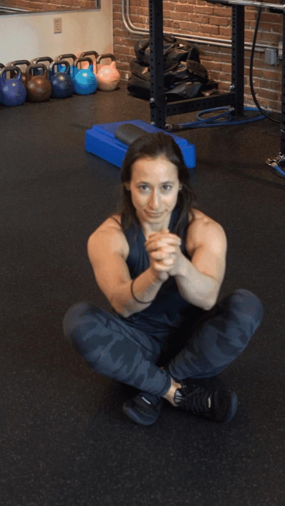 A woman in athletic wear sits cross-legged on a gym floor, hands clasped in front, with kettlebells and gym equipment behind her—capturing the focus and energy of Boston personal training.