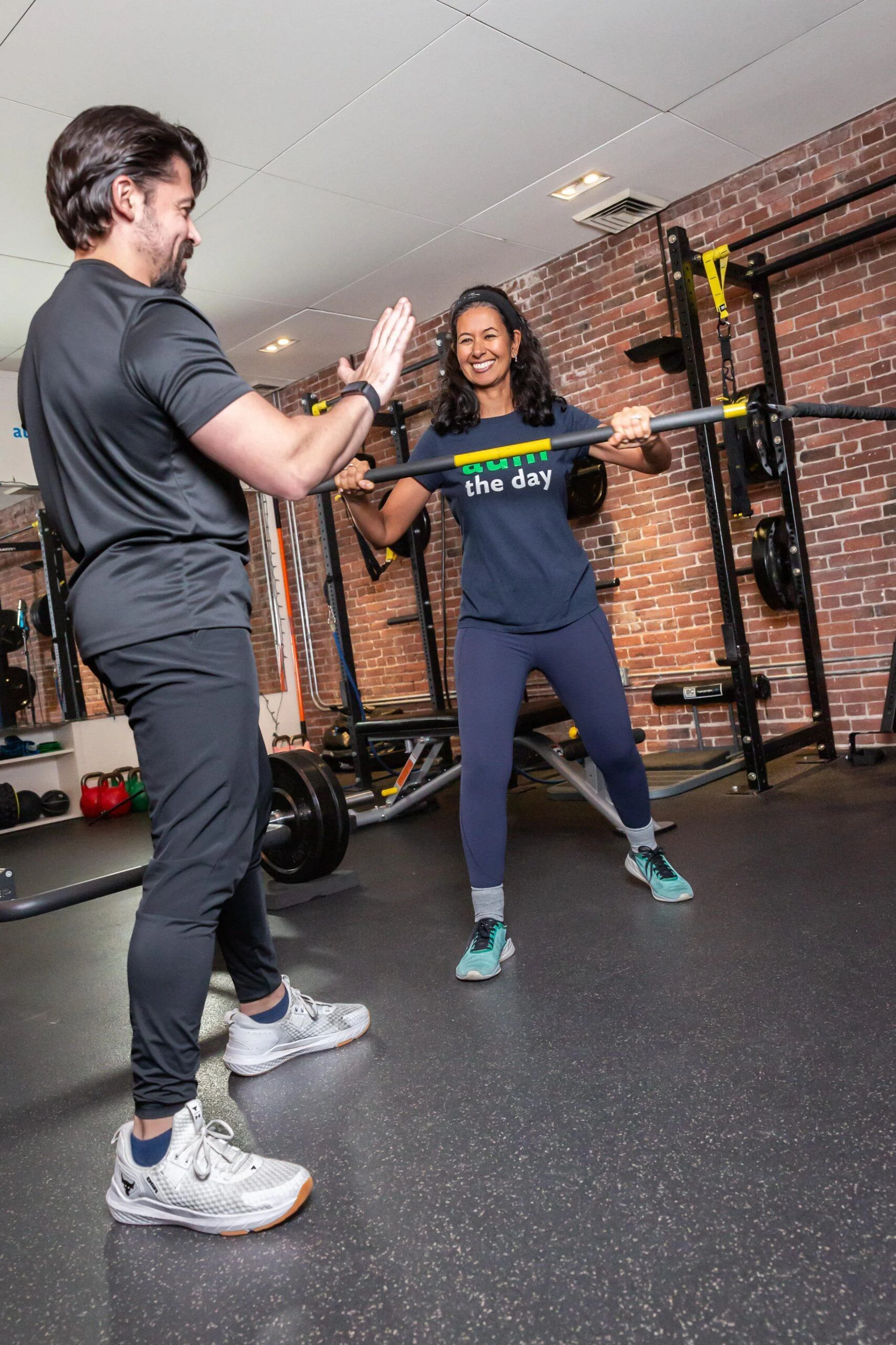 A woman lifts a barbell while smiling and giving a high-five to her male trainer in a gym with brick walls and workout equipment, inspiring others wondering how much does a personal trainer cost?