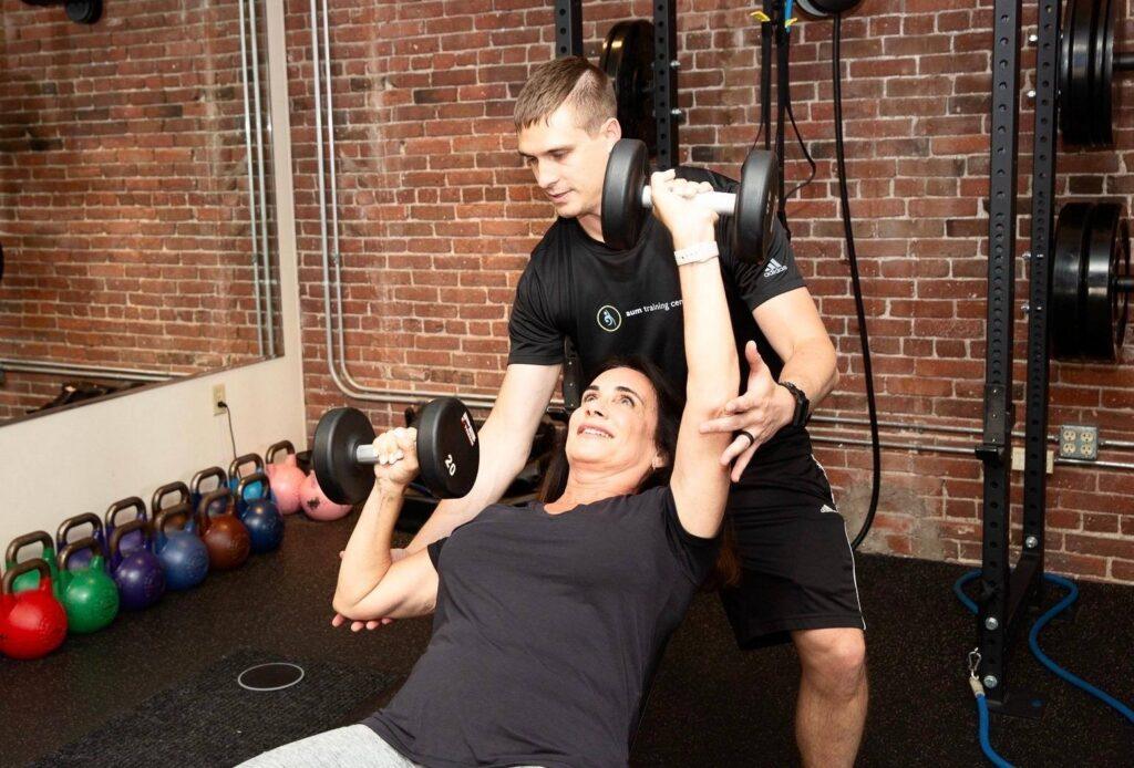 boston-personal-trainer-health About: A woman lifts dumbbells on a bench while a trainer assists her in a gym with kettlebells and weight racks in the background.