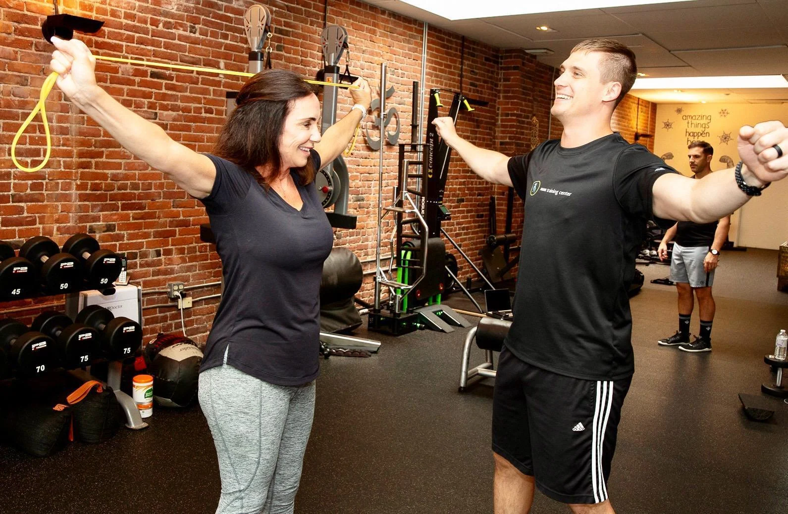 boston-personal-trainer-pricing A woman and a man exercise with resistance bands in a gym with brick walls and fitness equipment, reflecting the energetic vibe you’d find when exploring Boston personal trainer pricing options.