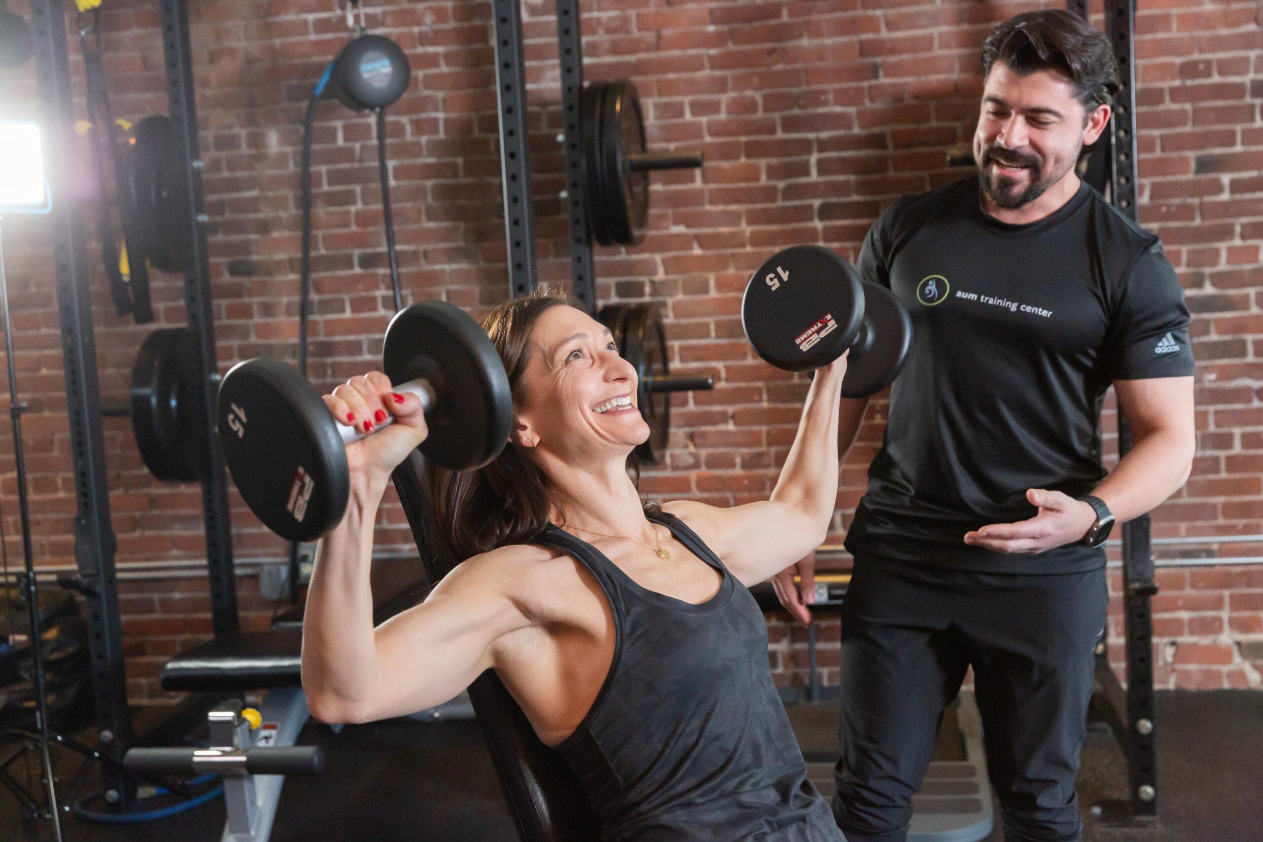 A woman performs a seated dumbbell shoulder press in a gym while a Boston personal trainer stands nearby, smiling.