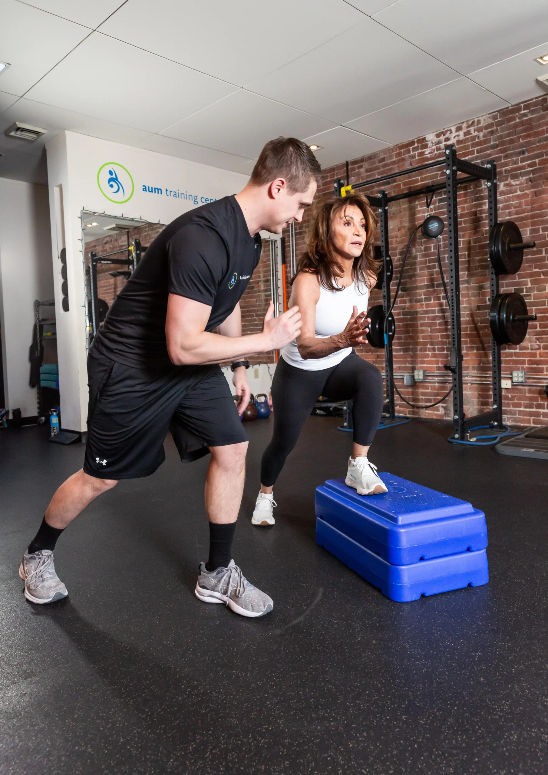 Media 1-88F3E03F-1B59-45A6-B2FE-CF1AB738440E A woman performs a step-up exercise on blue platforms while a trainer observes and instructs her in a gym with brick walls and fitness equipment.
