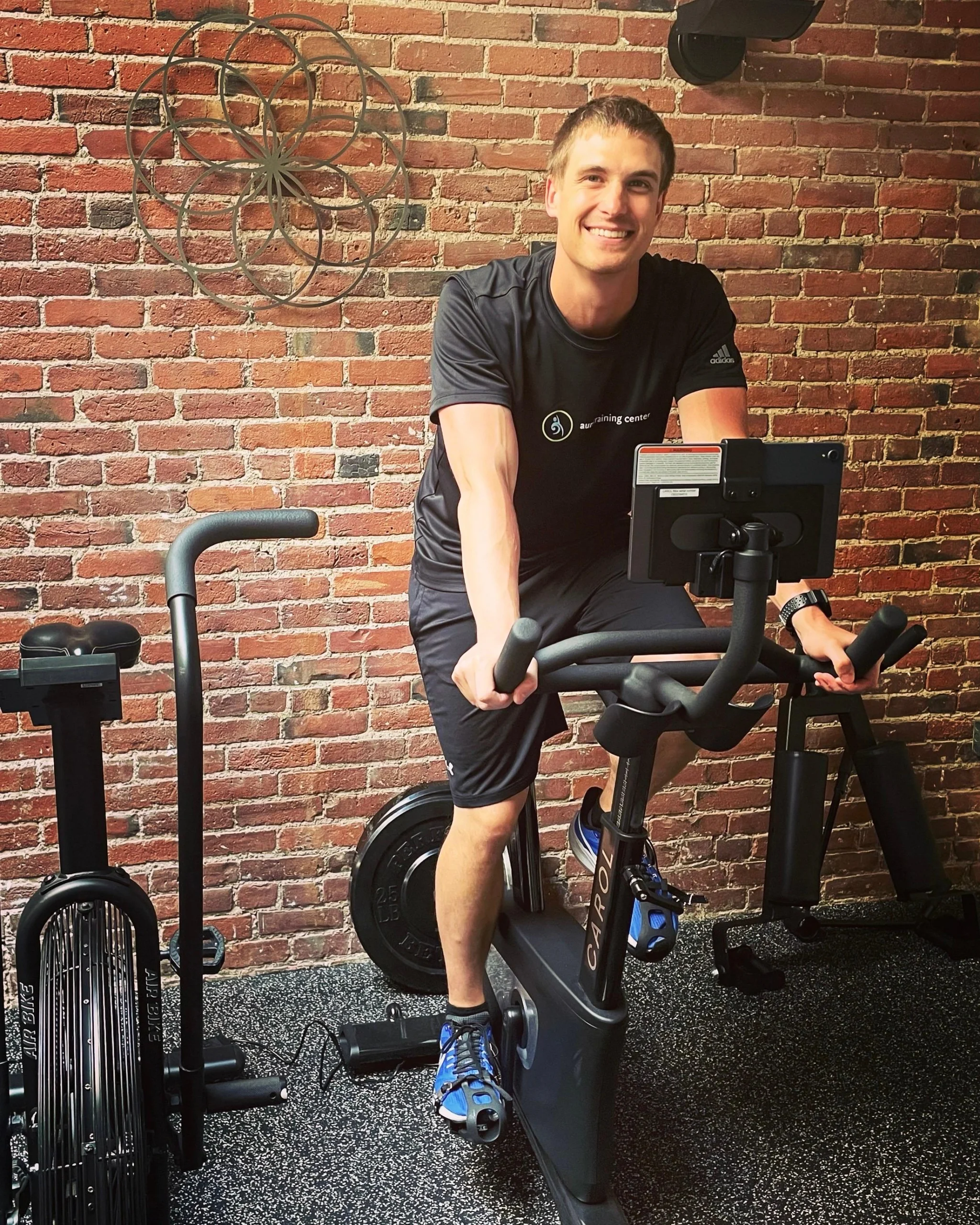 Media 2-17DDE576-FAB4-45F4-8533-44426623E5AD A man in athletic wear smiles while riding a stationary bike in a gym with exposed brick walls and exercise equipment.