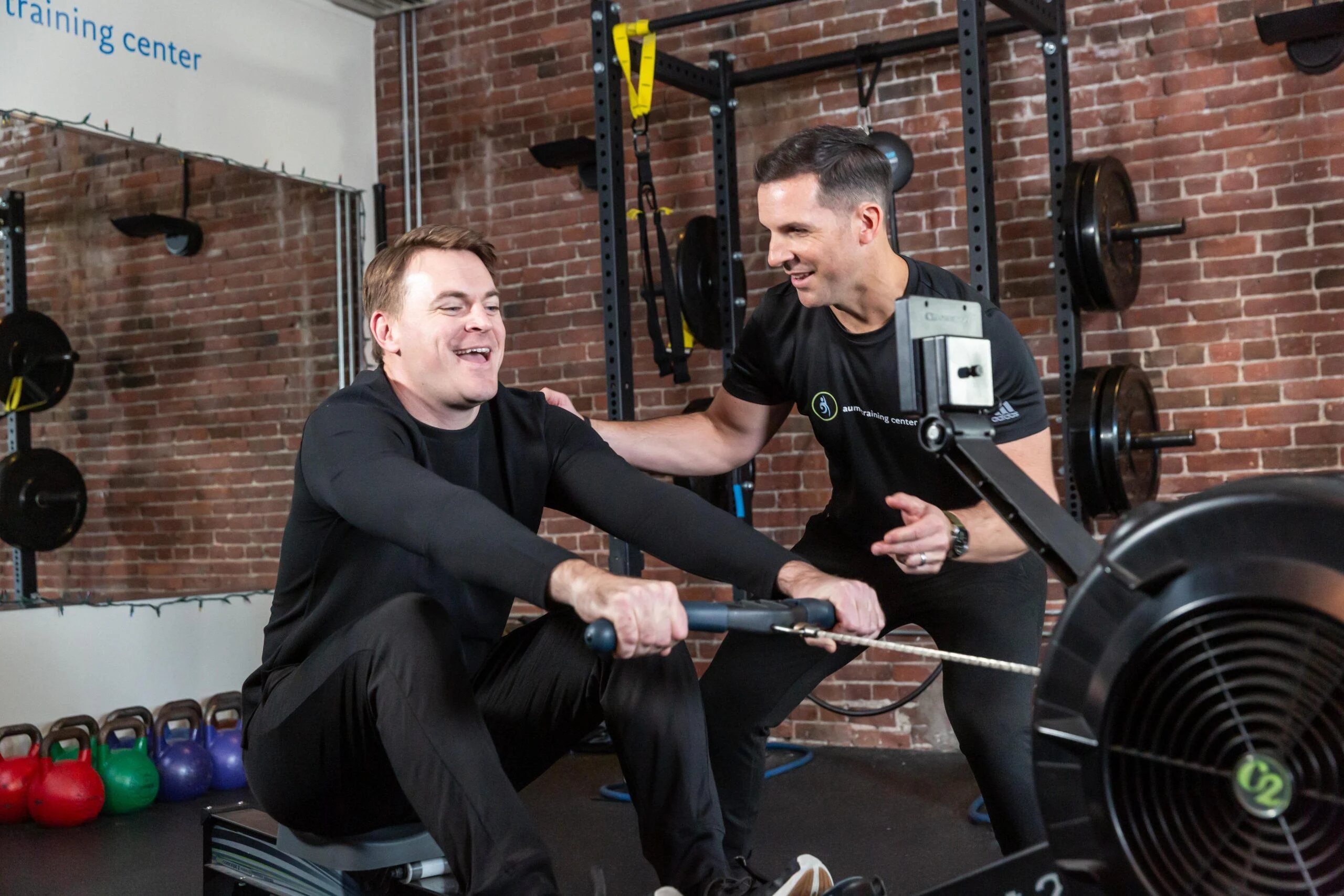 Man using a rowing machine while a trainer gives instructions and encouragement in a gym with weights, exercise equipment, and VO2 Max testing available in Boston.