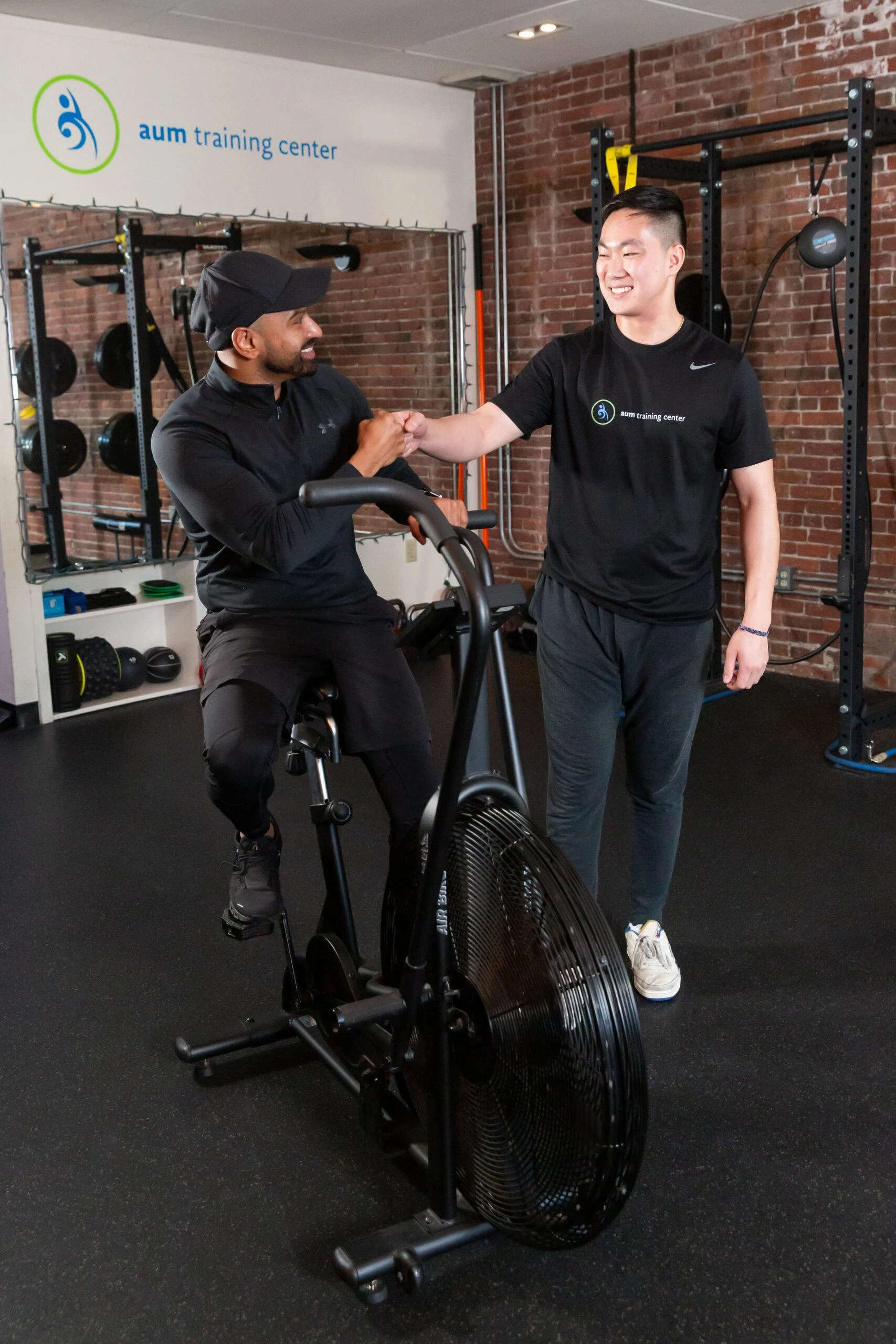 vo2-max-testing-in-boston A man on a stationary bike and another man standing next to him exchange a handshake in a gym setting, as VO2 Max testing in Boston takes place with workout equipment in the background.