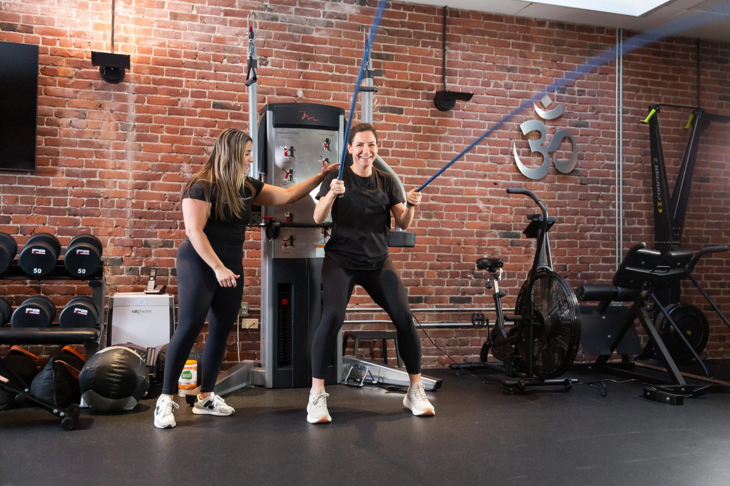 vo2-max-training-in-boston-aum-training Two women exercise in a gym; one pulls resistance bands while the other assists her. Both wear athletic clothes and stand in front of brick walls and gym equipment, highlighting an ideal space for VO2 Max testing in Boston.