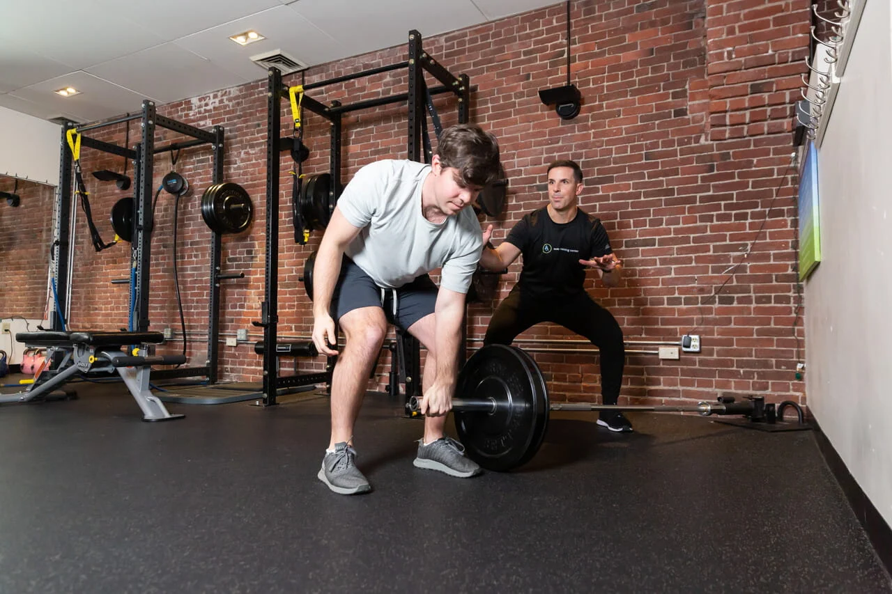 large-Media 1-86F0D1CD-6337-4302-98D1-E97A5AD4FA3A A man lifts a barbell in a gym while another man stands behind him, appearing to supervise or coach, highlighting the importance of safety and support during weightlifting sessions. Learn more about proper spotting techniques.