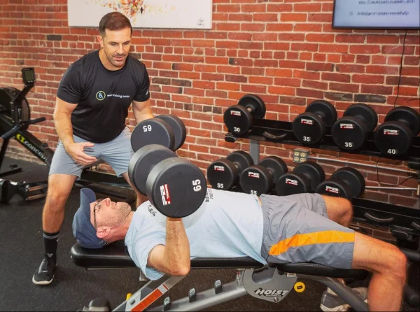 ‘Aum’ is a sacred sound that is known generally as the sound of the universe. Om is all encompassing, the essence of ultimate reality, and unifies everything in the universe. (1000 x 700 px) (600 (1) A man in a black shirt spots another man who is lying on a bench and lifting two 65 lb dumbbells at AUM Training Center, with a brick wall and weights in the background.