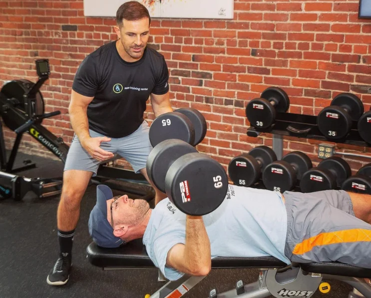 ‘Aum’ is a sacred sound that is known generally as the sound of the universe. Om is all encompassing, the essence of ultimate reality, and unifies everything in the universe. (1000 x 700 px) (600 (4) A man in a t-shirt and shorts observes another man doing a dumbbell bench press on a workout bench in a brick-walled gym, highlighting the benefits of onsite personal training.