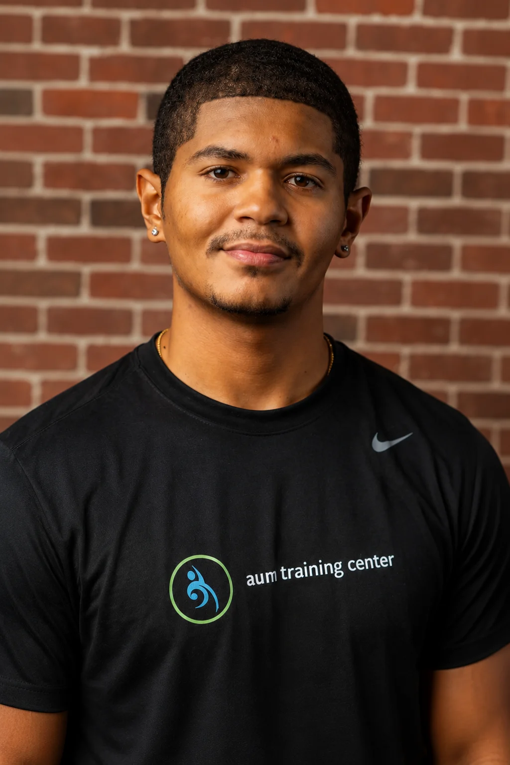 A man with short hair and a trimmed beard wearing a black "aum training center" T-shirt stands in front of a red brick wall.