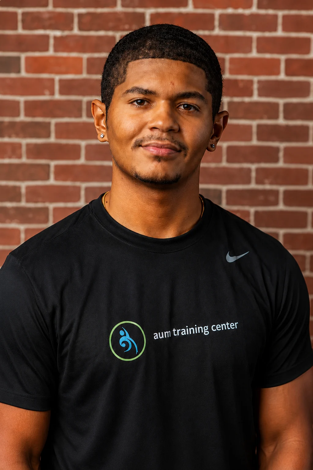 ChatGPT Image Jun 8, 2026, 09_13_23 AM A man stands in front of a brick wall, wearing a black "aum training center" t-shirt and looking at the camera with a neutral expression.
