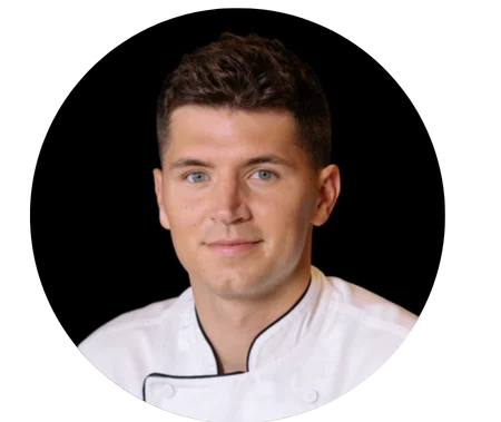 Untitled design – 2026-09-08T183744.440 A man with short brown hair in a white chef’s coat is looking at the camera against a black background, exuding the confident poise of a seasoned Boston Personal Trainer.